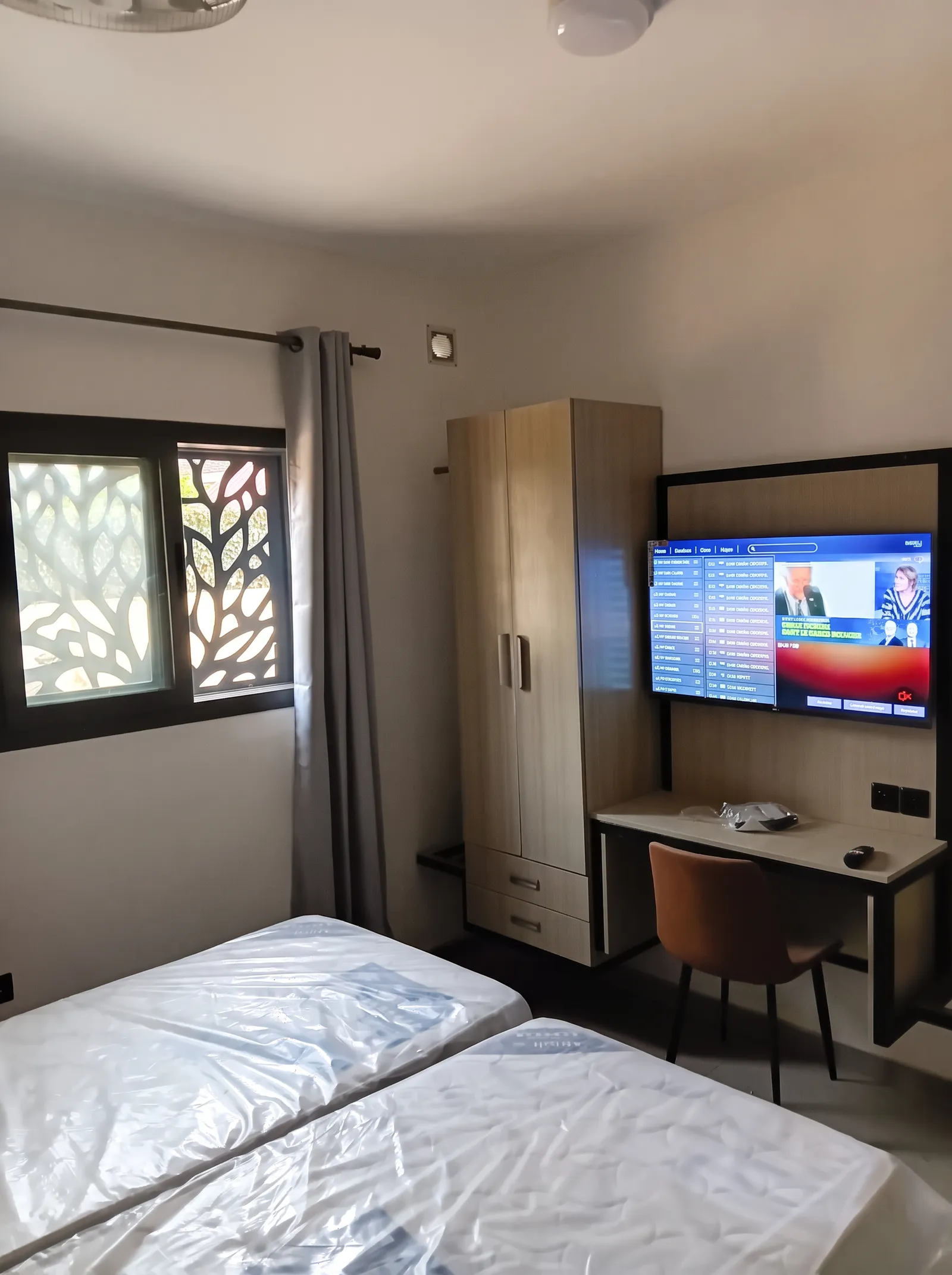 Chambre avec deux lits, bureau intégré, armoire et télévision murale, éclairée par une fenêtre à claustra.